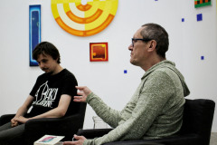 Sfotografowany z lewego profilu Robert Brzęcki siedzi w ciemnym fotelu. Za nim Piotr Tkacz z pochyloną głowa w ciemnej koszulce z białymi napisami. W głębi na białej ścianie wyeksponowane prace.