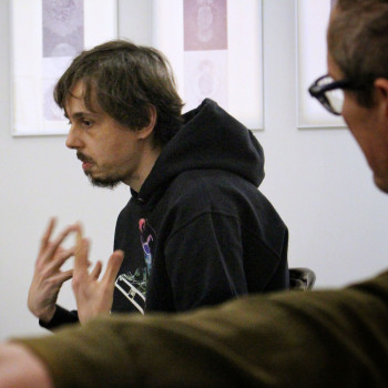 W centrum kadru Piotr Tkacz w ciemnej bluzie z kapturem. Gastykuluje wznosząc dłonie. Po prawej Robert Brzęcki sfotografowany z lewego boku.