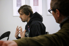 W centrum kadru Piotr Tkacz w ciemnej bluzie z kapturem. Gastykuluje wznosząc dłonie. Po prawej Robert Brzęcki sfotografowany z lewego boku.