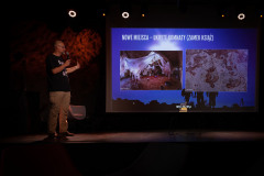 Tomasz Kulas na scenie wskazuje na slajd ze zdjęciami i napisem Nowe miejsca - ukryte komnaty (Zamek Książ).