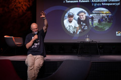 Tomasz Kulas siedzi na skraju sceny i podnosi w górę lewą rękę. W prawej trzyma mikrofon. W tle sladj z napisem i zdjęciem prowadzących spotkanie.