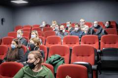 Sześć rzędów czerwonych foteli w sali kinowej. Zasiadają w nich widzowie w maseczkach. W pierwszym rzędzie kobieta w zielonej bluzce i czarnej maseczce.