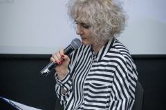 Paulina Pachulska-Wojdak w prawej dłoni trzyma mikrofon, w lewej notatki, z których czyta. Ubrana w koszulową bluzkę w białe i czarne pasy.