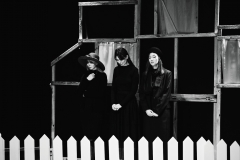 Trzy odziane w długie czarne suknie kobiety stoją rzędem przed białym płotkiem. Mają smutne twarze.