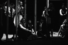 Grupa aktorów i aktorek ubranych w koszule z kołnierzykami. Stoją i siedzą pomiędzy słupami rozstawionymi na scenie.