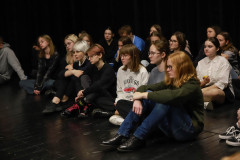 Młodzież siedząca na ciemnych deskach sceny.