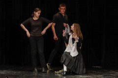 Trzy osoby w trakcie odgrywania scenki. Chłopak i dziewczyna w ciemnych spodniach i koszulkach stoją. Dziewczyna w ciemnej bluzce, plisowanej spódnicy i białej apaszce siedzi.