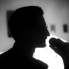 Ciemny zarys postaci sfotografowanej do ramion. Mężczyzna sfotografowany z prawego profilu. W dłoni trzyma mikrofon zbliżony do ust. Zdjęcie czarno-białe. 