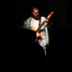 Mrok a w centrum kadru oświetlona postać gitarzysty. 