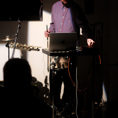 Mężczyzna stojący przy instrumentach perkusyjnych w świetle i mroku. Po lewej ciemna sylwetka. 