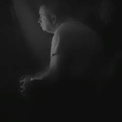 W centrum kadru rozmyta postać siedzącego mężczyzny w jasnej koszulce. Mężczyzna sfotografowany z lewego profilu.  Zdjęcie czarno-białe.
