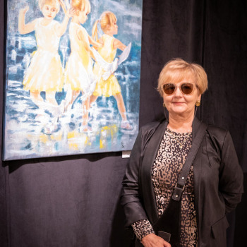 Kobieta w sukience we wzorki i ciemnym żakiecie pozuje do zdjęcia na tle pracy przedstawiającej trzy tańczące dziewczynki w żółtych sukienkach.