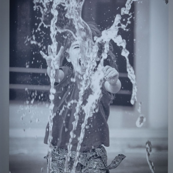 Zbliżenie na jedną z prac. Zdjęcie roześmianego dziecka próbującego chwycić strumień fontanny.