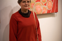 Kobieta w czerwonej bluzce i okularach pozuje na tle jednej z prac.