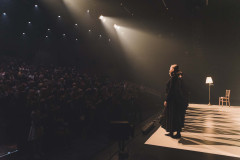 Rząd kobiet w ciemnych sukniach stoi na brzegu sceny. Nad pogrążoną w półmroku stojąca publicznością punktowe światła i trzy smugi reflektorów.