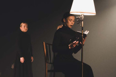 Za zapaloną stojącą lampą siedzi kobieta czytająca książkę. W głębi kadru wyprostowana tancerka w długiej ciemnej sukience.