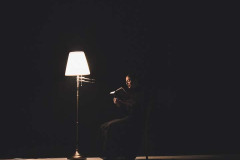 Kobieta siedzi na krześle na scenie. Czyta książkę. Obok niej zapalona lampa.