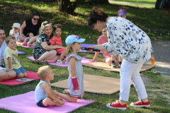 Kobiety i dzieci siedzą na matach na trawie. Na pierwszym planie kobieta przybliża mikrofon do ust klęczącej przed nią dziewczynki.