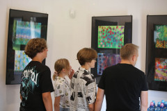 Cztery osoby odwrócone plecami do obiektywu oglądają dziecięce prace w antyramach wyeksponowane na ścianie.