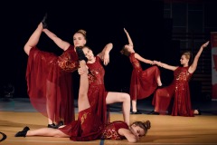Pięć młodych dziewczyn w czerwonych sukniach wykonuje figury taneczne na scenie.