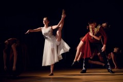 Młoda dziewczyna w białej sukni podnosi lewą nogę wysoko do góry. Po prawej stronie chłopak w czerwonym stroju unosi na plecach tancerkę.