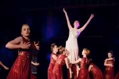 Grupa młodych dziewczyn w czerwonych sukniach ze złotymi elementami unosi tancerkę ubraną w białą sukienkę falującą u spodu. Po lewej stronie tancerka.