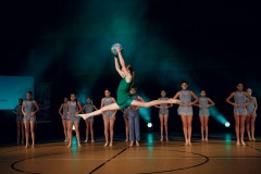 Dziewczyna w krótkiej zielonej sukience wykonuje w powietrzu szpagat bokiem do publiczności. Ponad głową trzyma niebieską kulę. Z nią widać tancerki ubrane na błękitno.