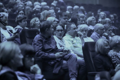Publiczność zgromadzona w sali, zasiadająca w fotelach. Zdjęcie wykonane z użyciem szarego filtra.