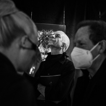 Na pierwszym rozmytym planie rozmawia ze sobą dwoje ludzi. W tle pomiędzy nimi Tadeusz Topolski stoi z uniesioną głową. Zdjęcie czarno-białe.