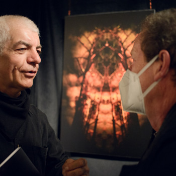 Tadeusz Topolski rozmawia z mężczyzną w białej maseczce. W tle zawieszone pod sufitem zdjęcie artysty. Przedstawia ono czarne konary drzew otoczone czerwonymi obłokami.