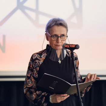 Krystyna Mikołajczak stoi przed mikrofonem. W dłoniach trzyma teczkę. Za nią widać slajd z logiem Centrum Kultury i Sztuki w Koninie.