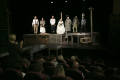 Aktorzy spektaklu Kyś stoją rzędem na scenie.  Zdjęcie wykonane z widowni.