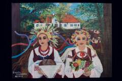 Obraz przedstawiający dwie młode kobiety w strojach ludowych. Kobieta po lewej stronie trzyma tacę z bochenkiem chleba. Natomiast pani po prawej kosz owoców. Za nimi widać biały dworek z czerwonym dachem.