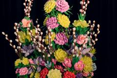 Stroik wielkanocny złożony z różnokolorowych kwiatów. Kwiaty wykonane są z bibuł. Pomiędzy umieszczone bazie.