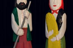 Dwie figury drewniane. Po lewej stronie mężczyzna z brodą, w niebieskiej wysokiej czapce  trzyma kosę. Po prawej kobieta z czerwoną chustą na głowie trzyma zboże. Oboje ubrani są w stroje ludowe.
