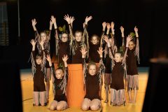 Grupa 10 dziewczynek w brązowych strojach z frędzlami unosi w górę ręce.