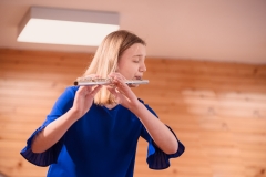 Dziewczyna w chabrowej sukience z rozkloszowanymi rękawami gra na flecie.