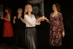 Kobieta z blond włosami z długimi ciemnymi kolczykami, w białej bluzce i ciemnej spódni niesie w dłoniach lampion. W tle trzy kobiety ze świeczkami.