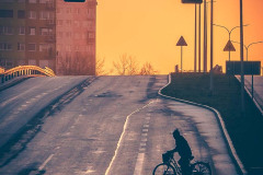 Wiadukt. Na pierwszym planie ciemna sylwetka przejeżdżającego po pasach rowerzysty. Za garbem wiaduktu dwa wieżowce i pomarańczowe niebo.