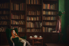 Rudowłosa młoda kbieta siedzi w zielonym fotelu z rozrzuconymi nogami. W tle biblioteczka wypełniona książkami.