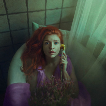 Kobieta w liliowej sukience leży w wannie ze słuchawką starego telefonu. Patrzy w obiektyw. Po prawej zwoje tiulu.