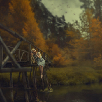 Kobieta siedzi na drewnianym moście. W tle jesienne drzewa.