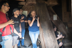 Grupa osób z aparatami fotograficznymi przy drewnianej konstrukcji wewnątrz wiatraka.