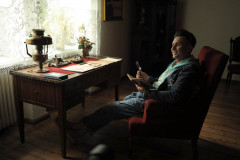 Prowadzący warsztaty wycięgnięty w czerwonym fotelu z książką i lupą. Przed nim biurko z dokumentami i stylową lampą.