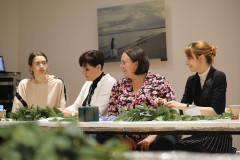 Cztery uczestniczki warsztatów siedzą przy stole. Na blacie rozłożone gałązki i cztery zielone świeczki w foliowym opakwoaniu. W tle na ścianie wisi praca,