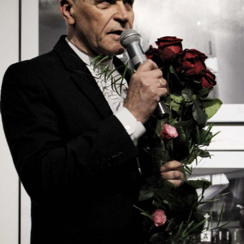 Franciszek Kupczyk z bukietem róż. W prawej dłoni trzyma mikrofon. Zdjęcie w planie amerykańskim.