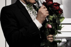 Franciszek Kupczyk z bukietem róż. W prawej dłoni trzyma mikrofon. Zdjęcie w planie amerykańskim.