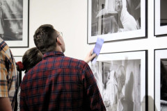Mężczyzna w czerwonej koszuli w kratkę ogląda prace na ścianie. Zasłania stojącą za nim kobietę, która wskazuje dłonią na zdjęcie.