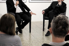 W fotelach na podwyższeniu siedzą Franciszek Kupczyk i Robert Brzęcki. Fotograf trzyma mikrofon. Na pierwszym planie tył głowy kobiety i mężczyzny.