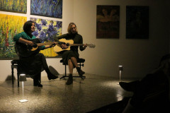 Dwie grające na gitarach kobiety siedzą na taboretach. Za ich plecami wiszące na ścianie obrazy. Na podłodze dwie lampki. W prawym dolnym rogu zdjęcia wyświetlacz telefonu.